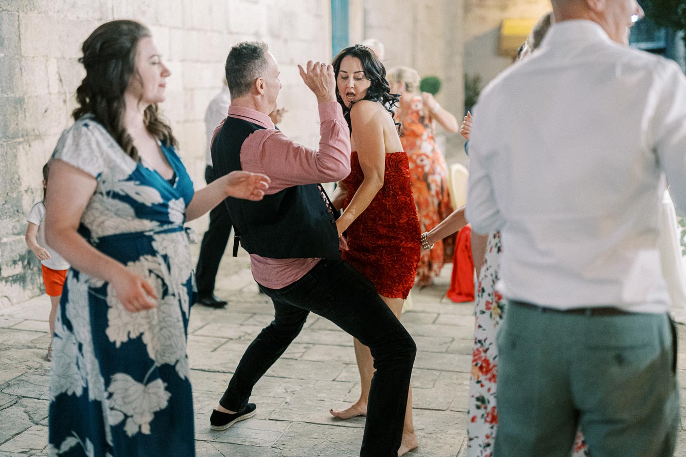 A group of people dancing energetically at a wedding reception, featuring a woman in a red dress and a man in a pink shirt with a vest, amidst a lively atmosphere with smiles and movement in an outdoor setting.