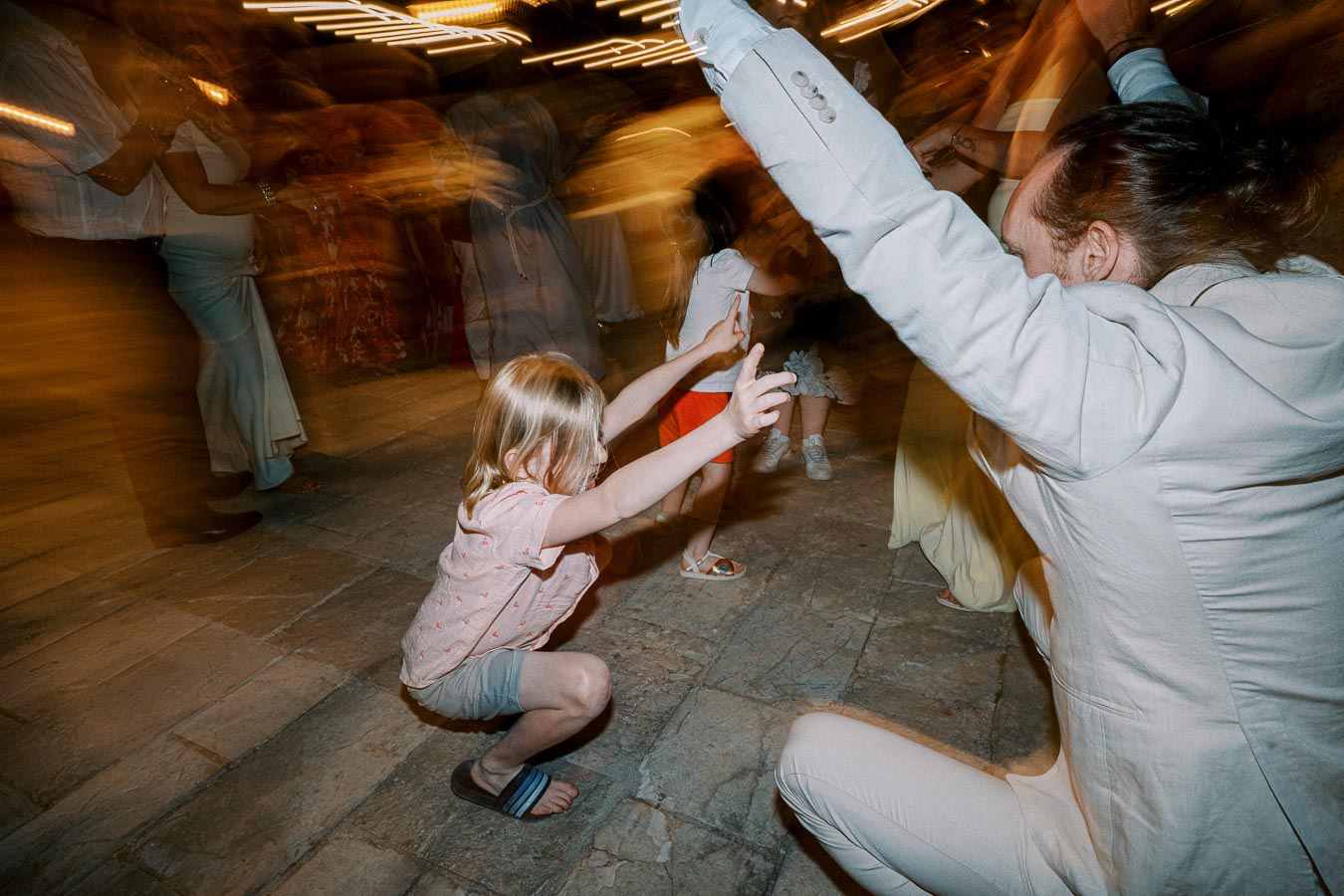 Children and adults dancing energetically at a lively evening event, with blurred motion capturing the excitement and festive atmosphere.
