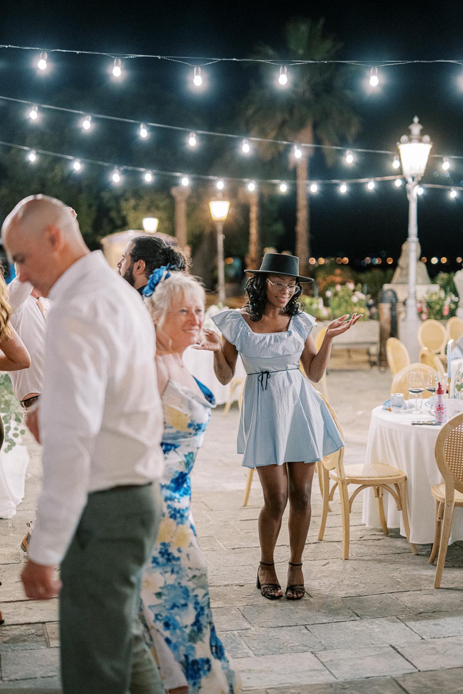 People enjoying an evening outdoor event under string lights, featuring a stylish woman in a light blue dress and hat, surrounded by other attendees in elegant attire.