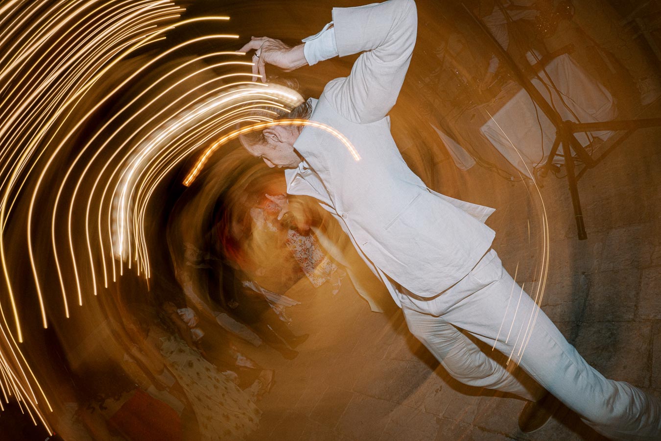 Man in a light suit dancing energetically at night with swirling light trails surrounding him, creating a dynamic and festive atmosphere.