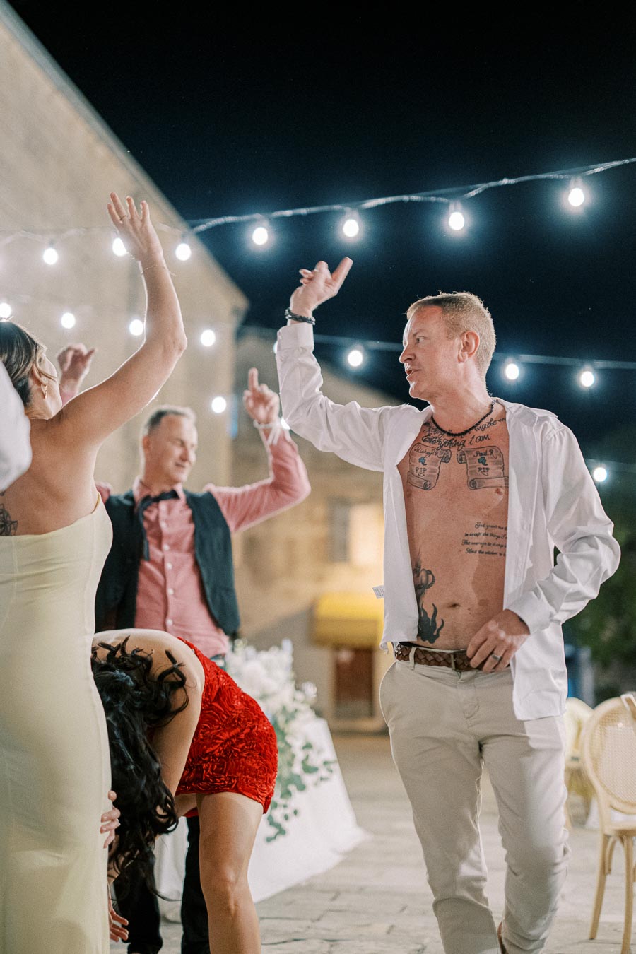 Group of people dancing energetically under string lights at an outdoor party, some wearing colorful dresses and white shirts, with a lively and joyful atmosphere.
