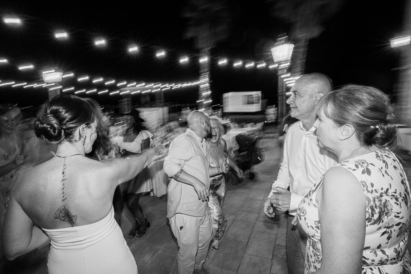 Black and white photograph of people dancing and laughing at an outdoor night event, with string lights creating a festive atmosphere.