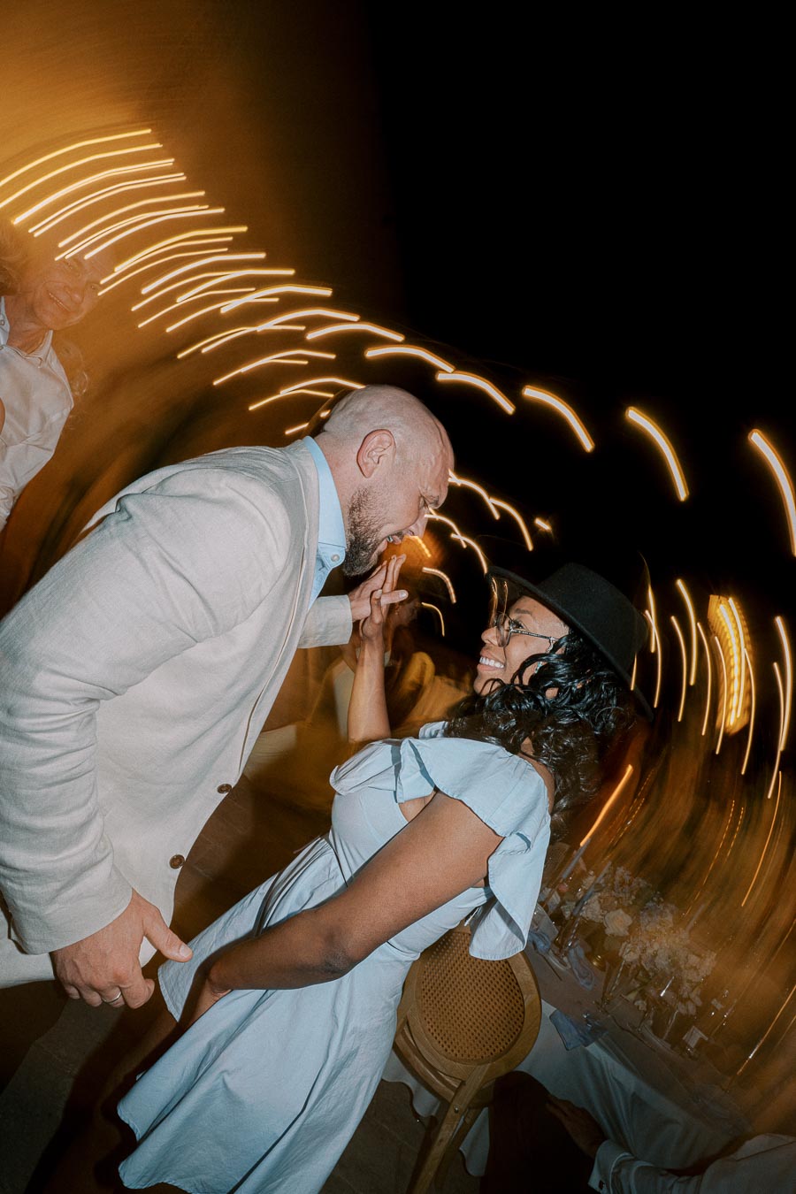 A happy couple dancing joyfully at a lively evening event, surrounded by creative light trails, with the man wearing a light suit and the woman in a blue dress and hat.