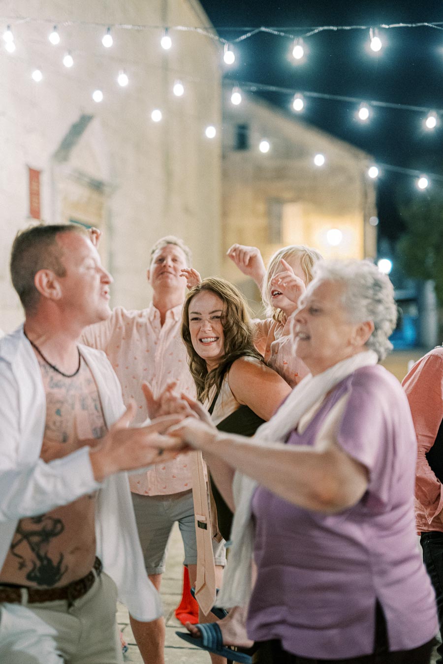 A group of people joyfully dancing under string lights at an outdoor evening celebration.