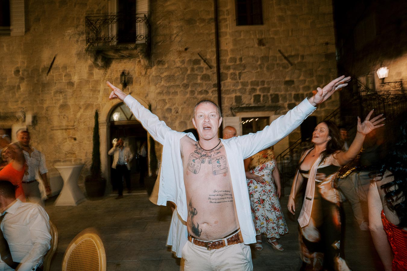 A joyful man with tattoos enthusiastically dances at an outdoor party, surrounded by other guests in stylish attire, against a backdrop of a rustic stone wall.