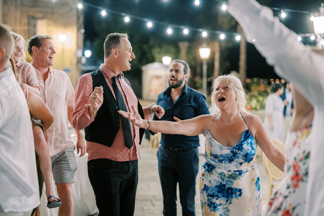 A group of people joyfully dancing and singing at an outdoor evening party, surrounded by string lights for ambiance.