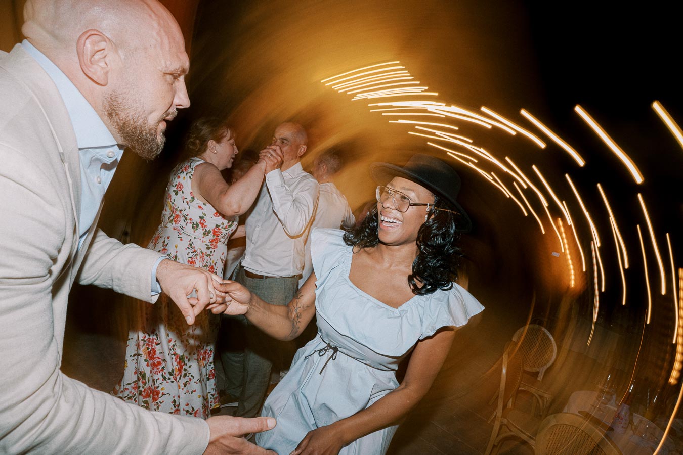 A joyful group of people dancing at a lively party, with a woman in a hat smiling brightly and warm lights creating a dynamic swirl effect in the background.