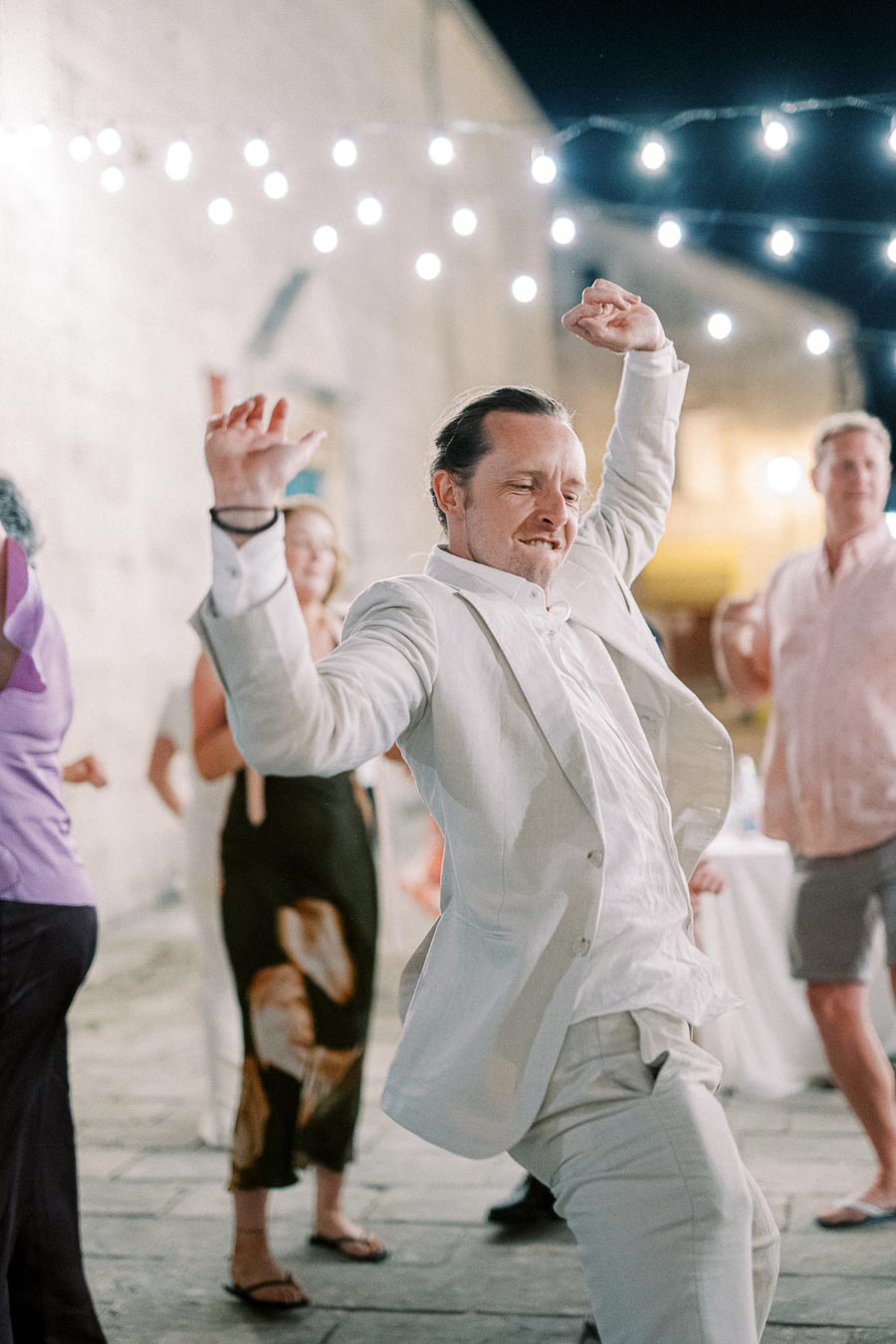 A man in a white suit energetically dancing outdoors under string lights at a lively evening event.