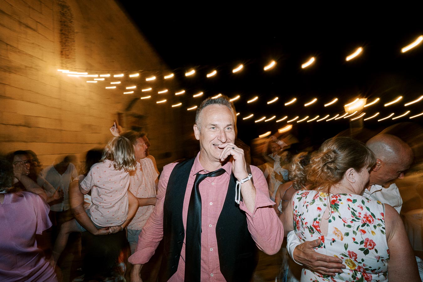 A joyful outdoor evening party with a man in a pink shirt and black tie laughing amidst dancing guests, under warm string lights.