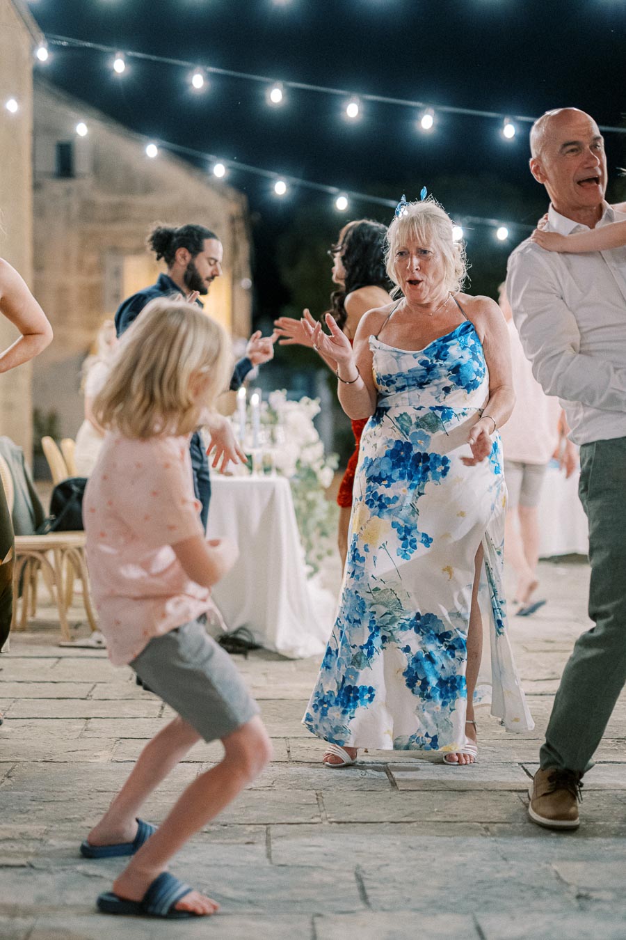 A group of people dancing joyfully under string lights at an outdoor evening event, with a woman in a blue floral dress leading the festivities.