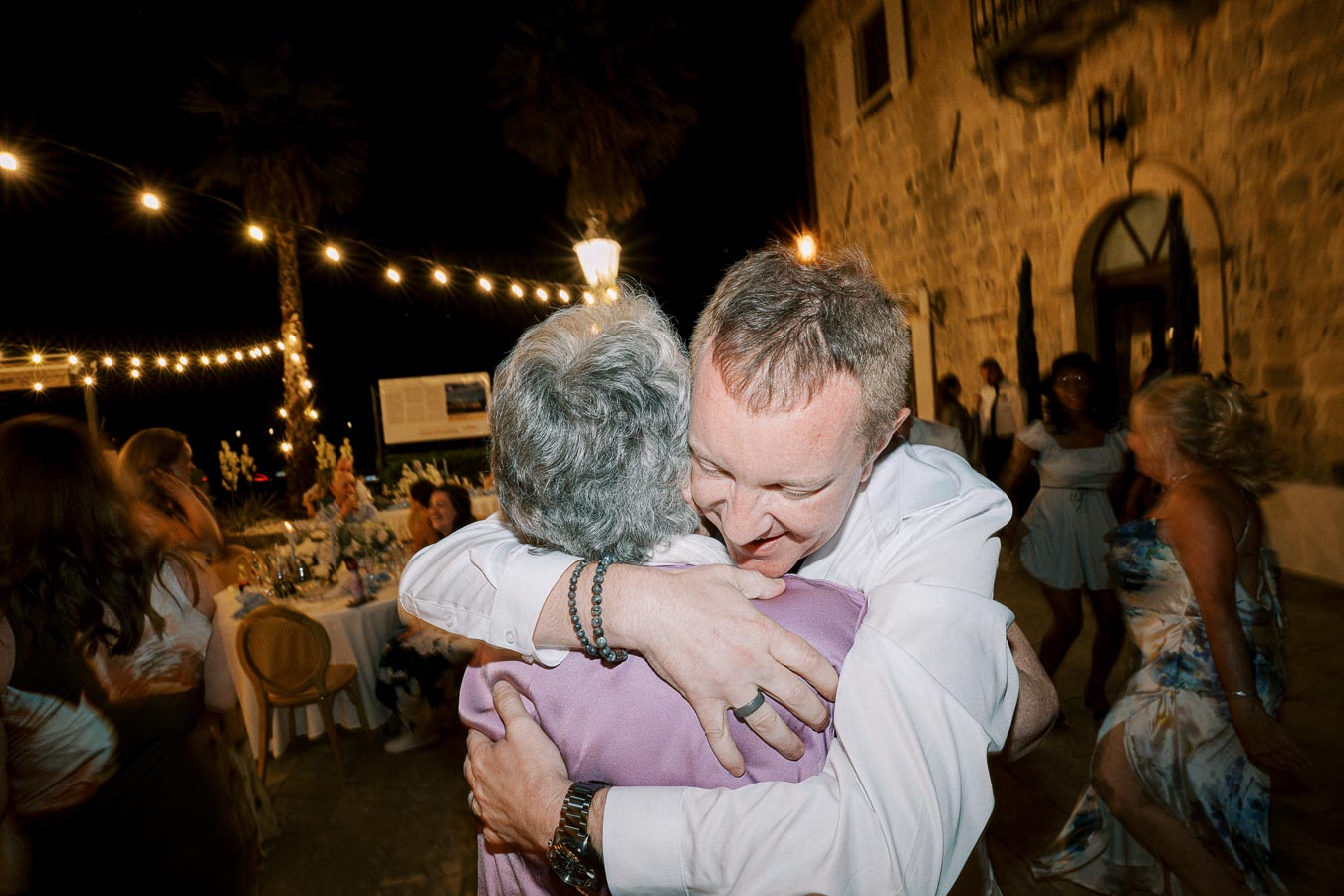 A joyous moment at an outdoor evening celebration, featuring two people embracing warmly. The scene is illuminated by string lights above, with a stone building visible in the background and several guests enjoying the festive atmosphere.