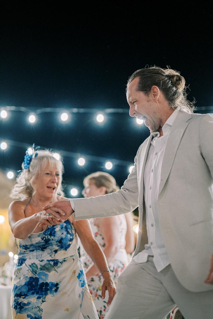 Elderly couple joyfully dancing under string lights at an outdoor evening event, both wearing floral-themed attire, exuding happiness and celebration.