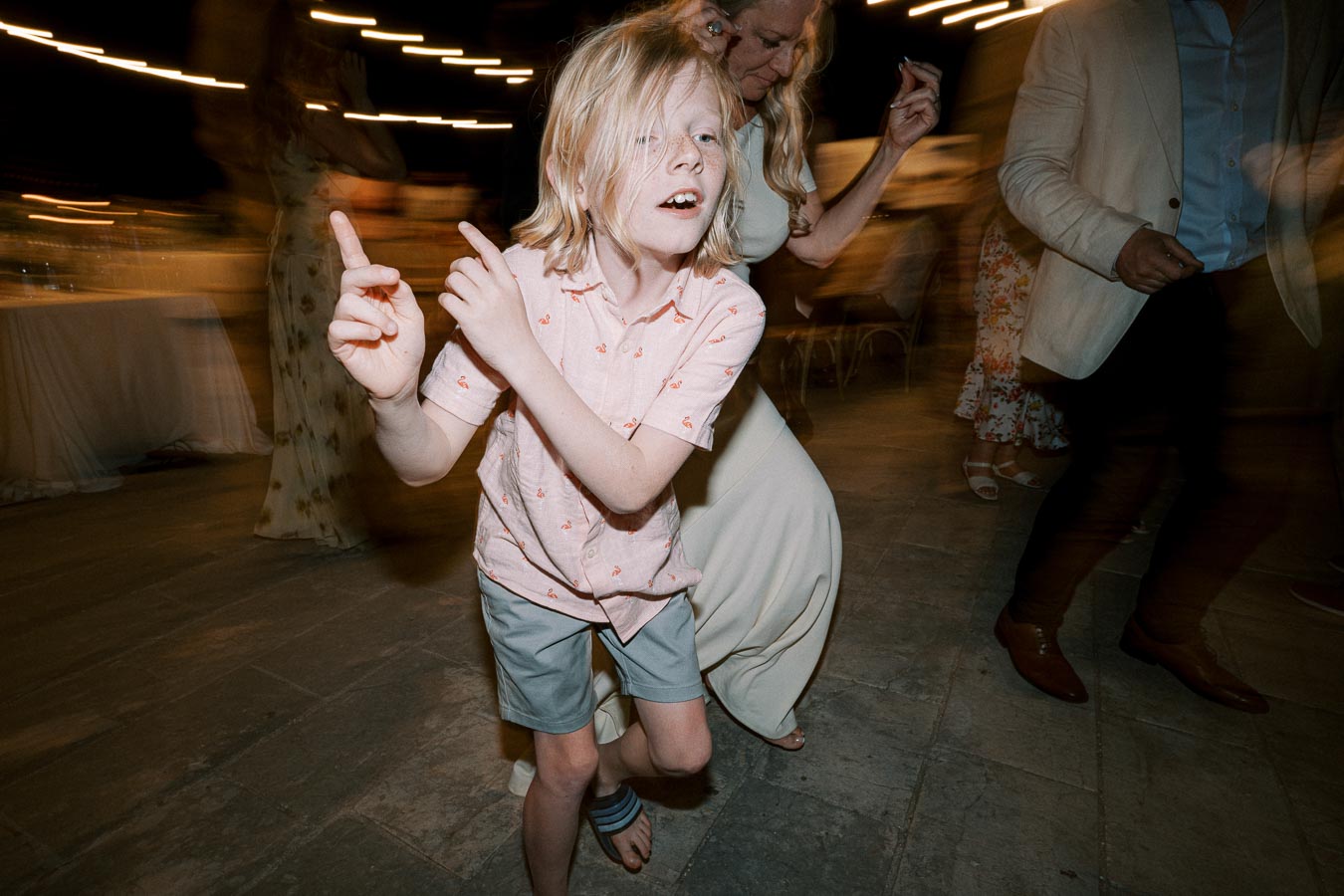 A young boy energetically dancing at a lively party, wearing a patterned shirt and shorts, surrounded by blurred motion of other partygoers in the background.