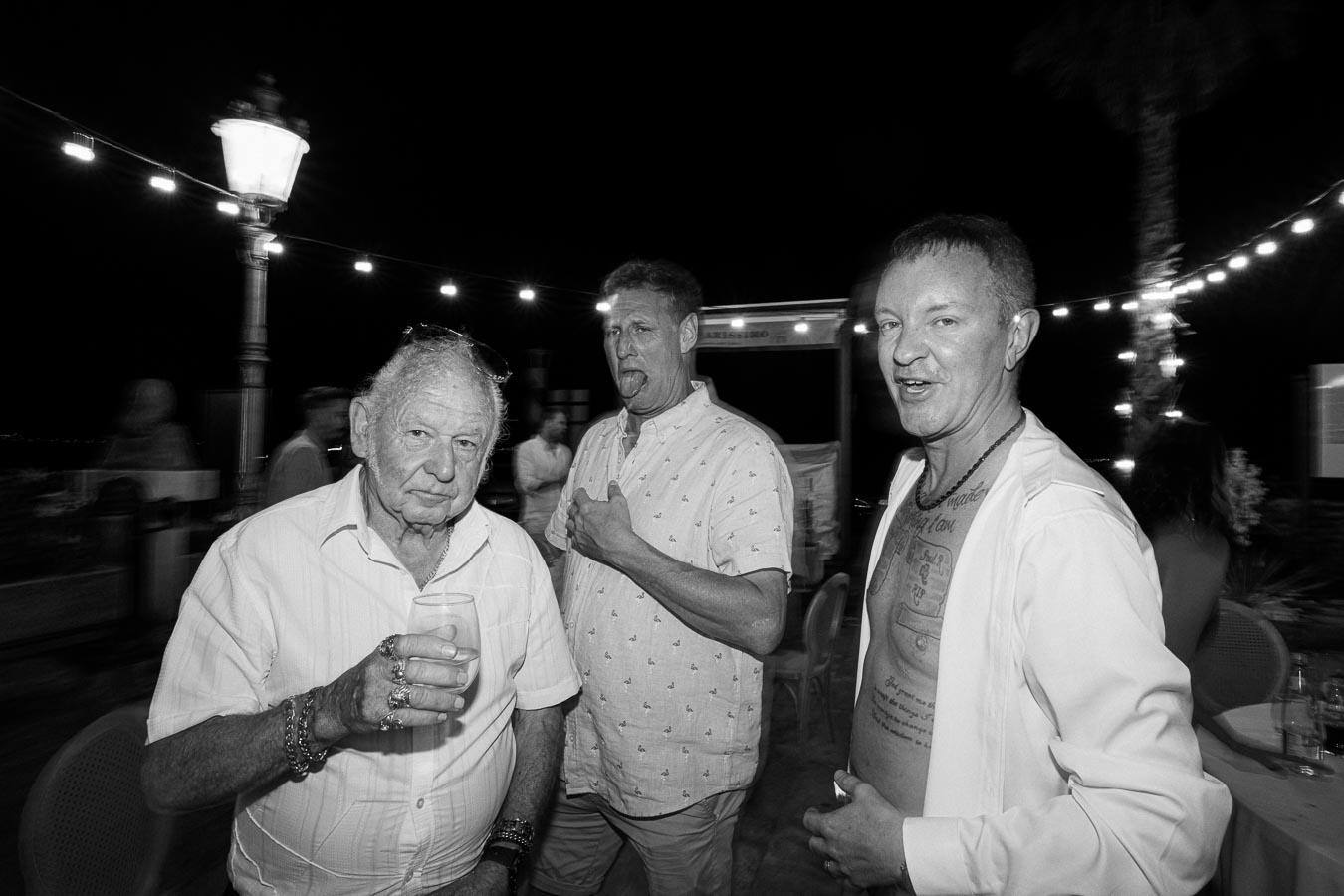 Black and white photo of three men at a night event, with one holding a drink, another making a funny face, and a third showing off tattoos, set against a backdrop of string lights and a lamppost.