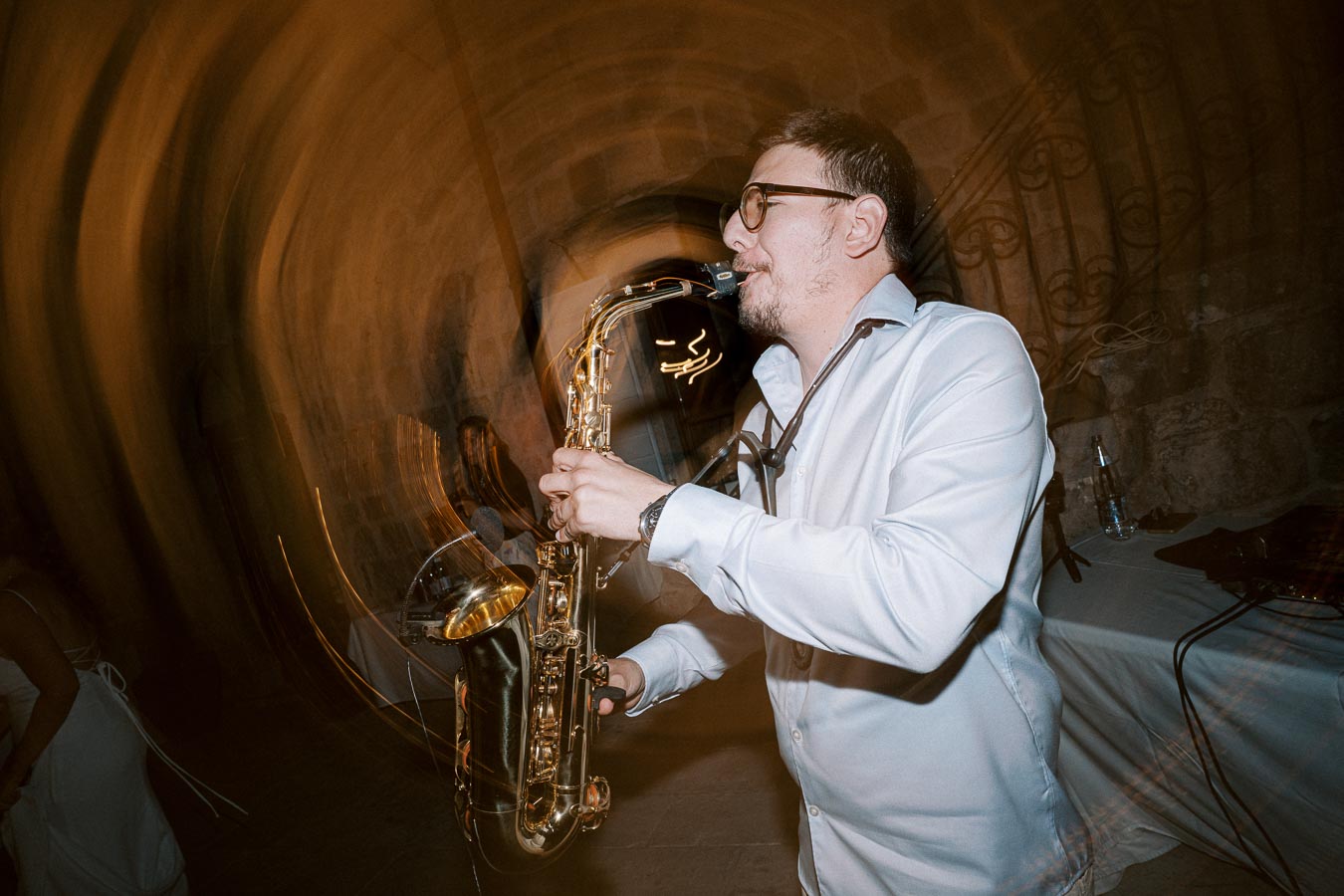 A musician playing a saxophone at an indoor event, wearing glasses and a white shirt. The background shows blurred motion effects and architectural details, suggesting movement and energy in a dimly lit setting.