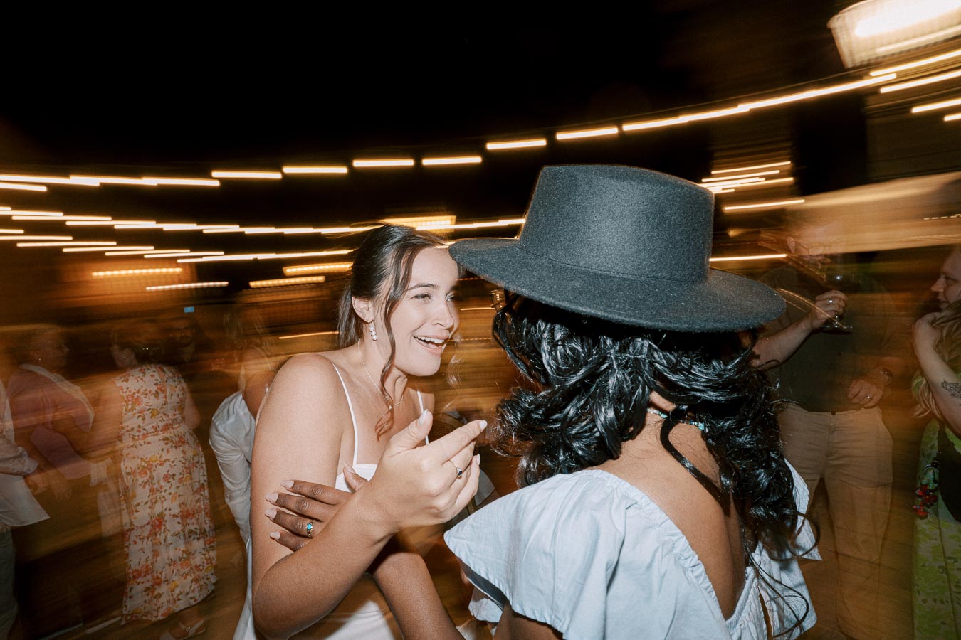 Women enjoying a lively conversation at a nighttime outdoor party, surrounded by blurred lights and a festive atmosphere.