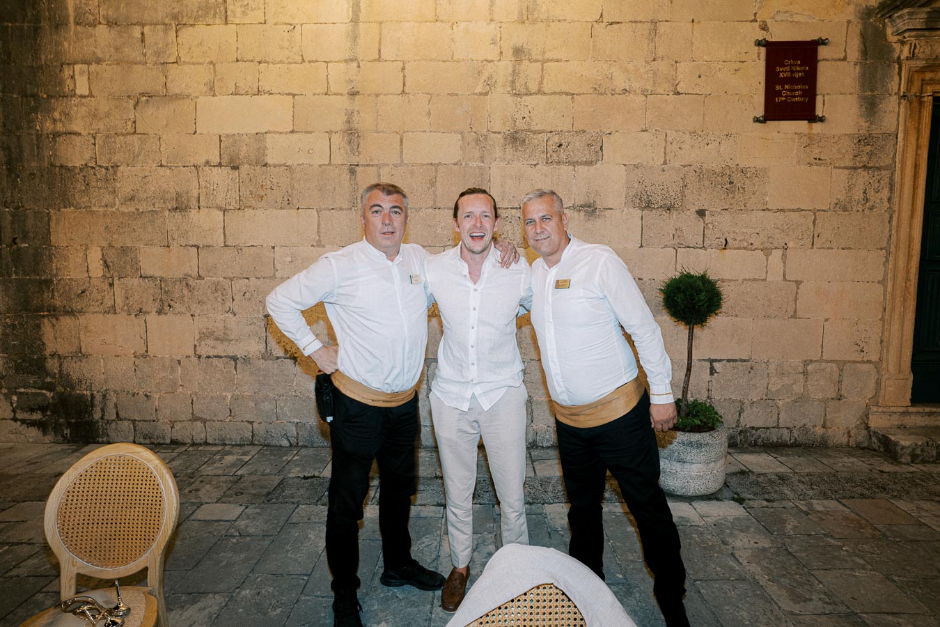 Three men smiling and posing together in front of a historic stone wall, dressed in white shirts. The setting includes a small potted plant and a wooden chair, suggesting an outdoor courtyard scene.