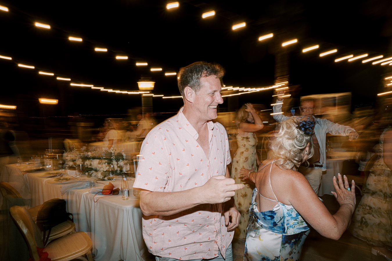 A joyful wedding reception dance scene at night, featuring guests smiling and wearing summer attire with blurred string lights in the background.