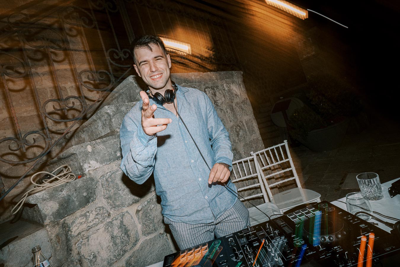 DJ wearing headphones smiling and pointing at the camera while standing behind a mixing console at an outdoor event.