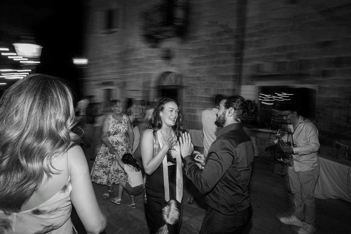 Black and white photo of a lively party scene with a couple joyfully dancing while others socialize and a musician plays the saxophone in the background.