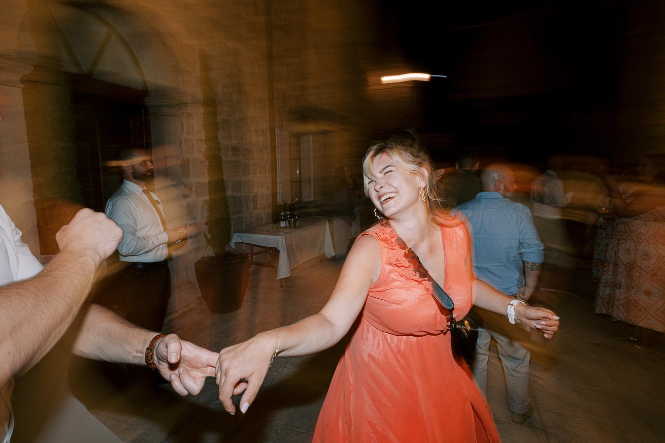 A woman in an orange dress smiling and dancing joyfully at a lively evening party, with blurred background of people and stone walls.