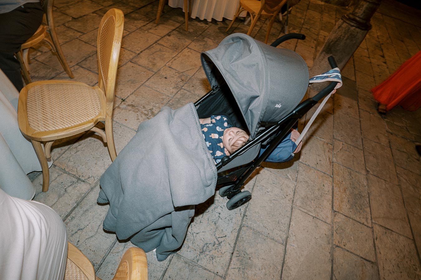 A sleeping baby in a stroller, covered with a gray blanket, surrounded by wooden chairs on a stone floor.