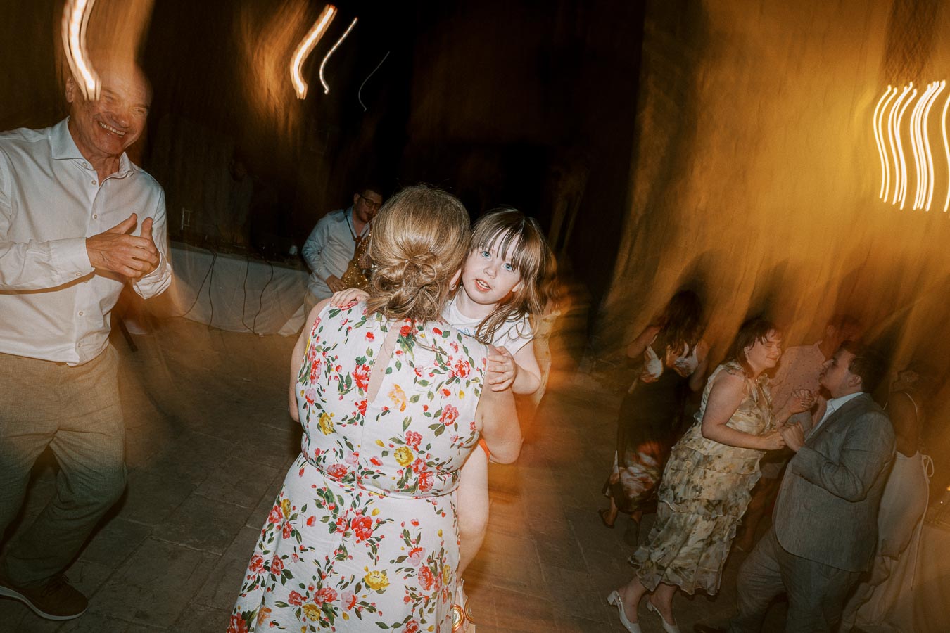 A joyful wedding reception dance floor scene with a woman in a floral dress carrying a child, surrounded by people dancing and celebrating under warm lighting.