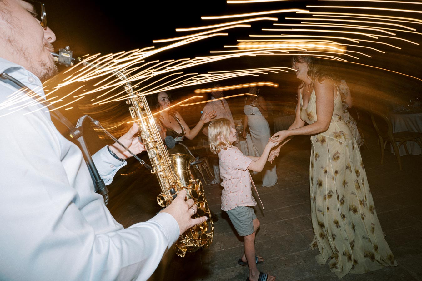 A saxophonist plays music at an outdoor event, with blurred lights creating dynamic patterns. A woman and child dance energetically in front of the musician, capturing a lively atmosphere.