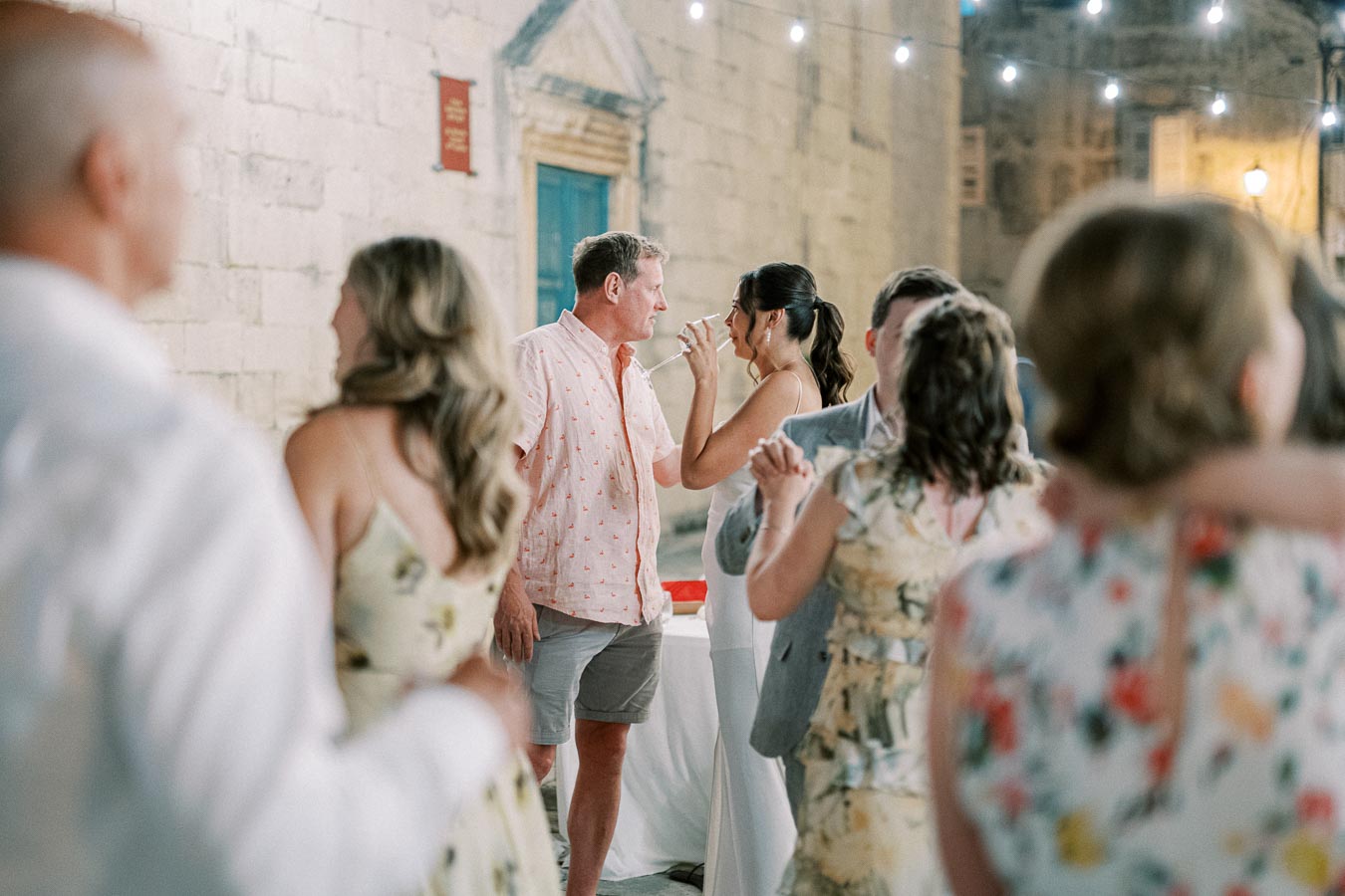 Outdoor evening celebration with people dancing and socializing under string lights near a stone building.