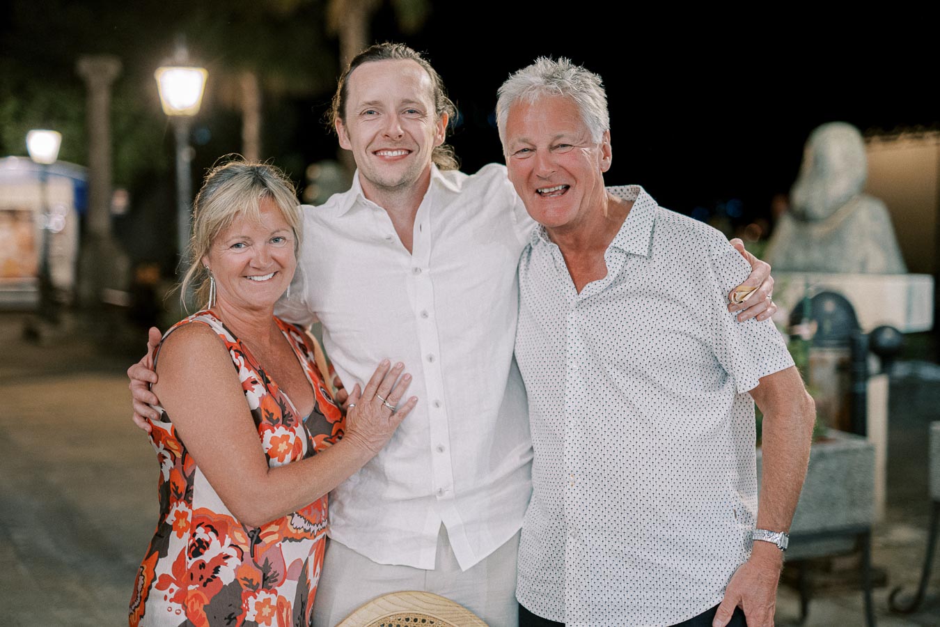 Three people smiling and embracing each other while enjoying an evening outdoors. The group stands under streetlights, with trees and night sky visible in the background, creating a warm atmosphere.
