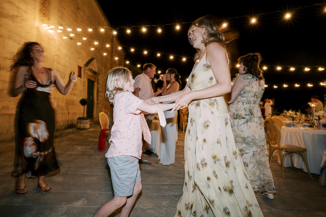 A joyful outdoor nighttime celebration with string lights overhead, where a woman in a floral dress dances with a young boy amidst other guests enjoying the festive atmosphere.
