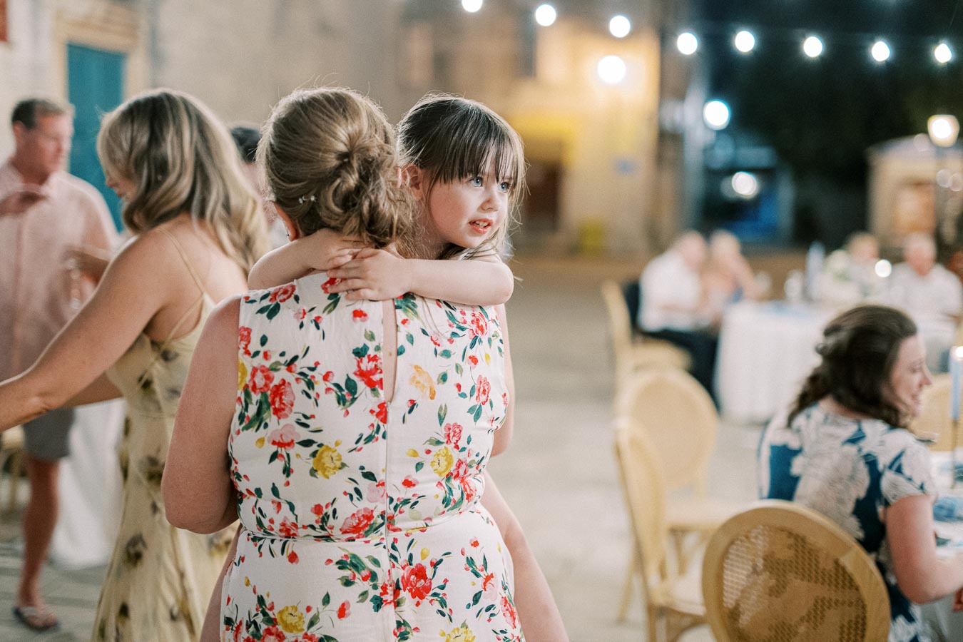 Outdoor evening gathering with string lights and tables, featuring a woman in a floral dress holding a young girl.