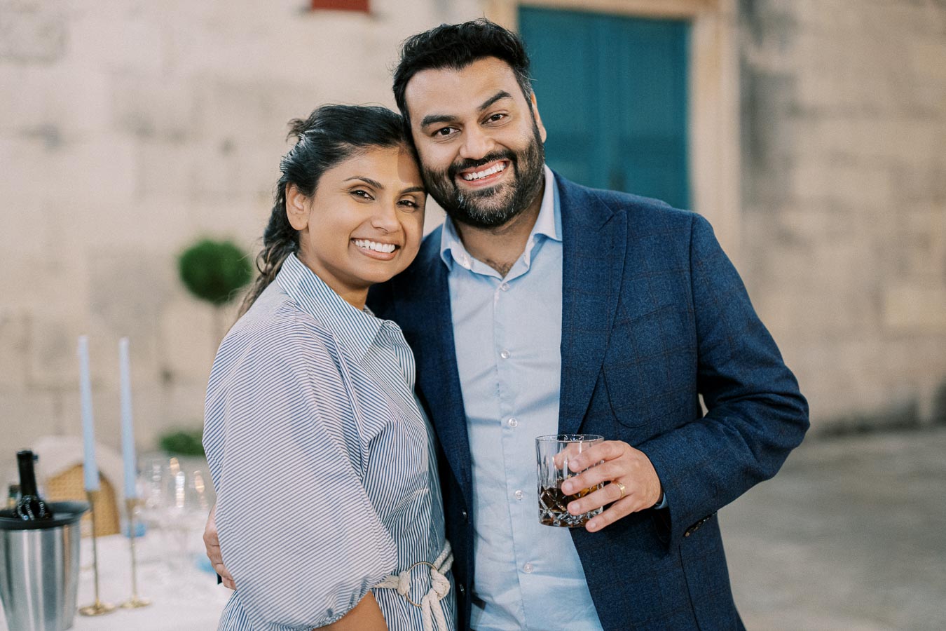 A smiling couple posing together at an outdoor event, with the man holding a drink and both dressed in smart casual attire against a blurred background.