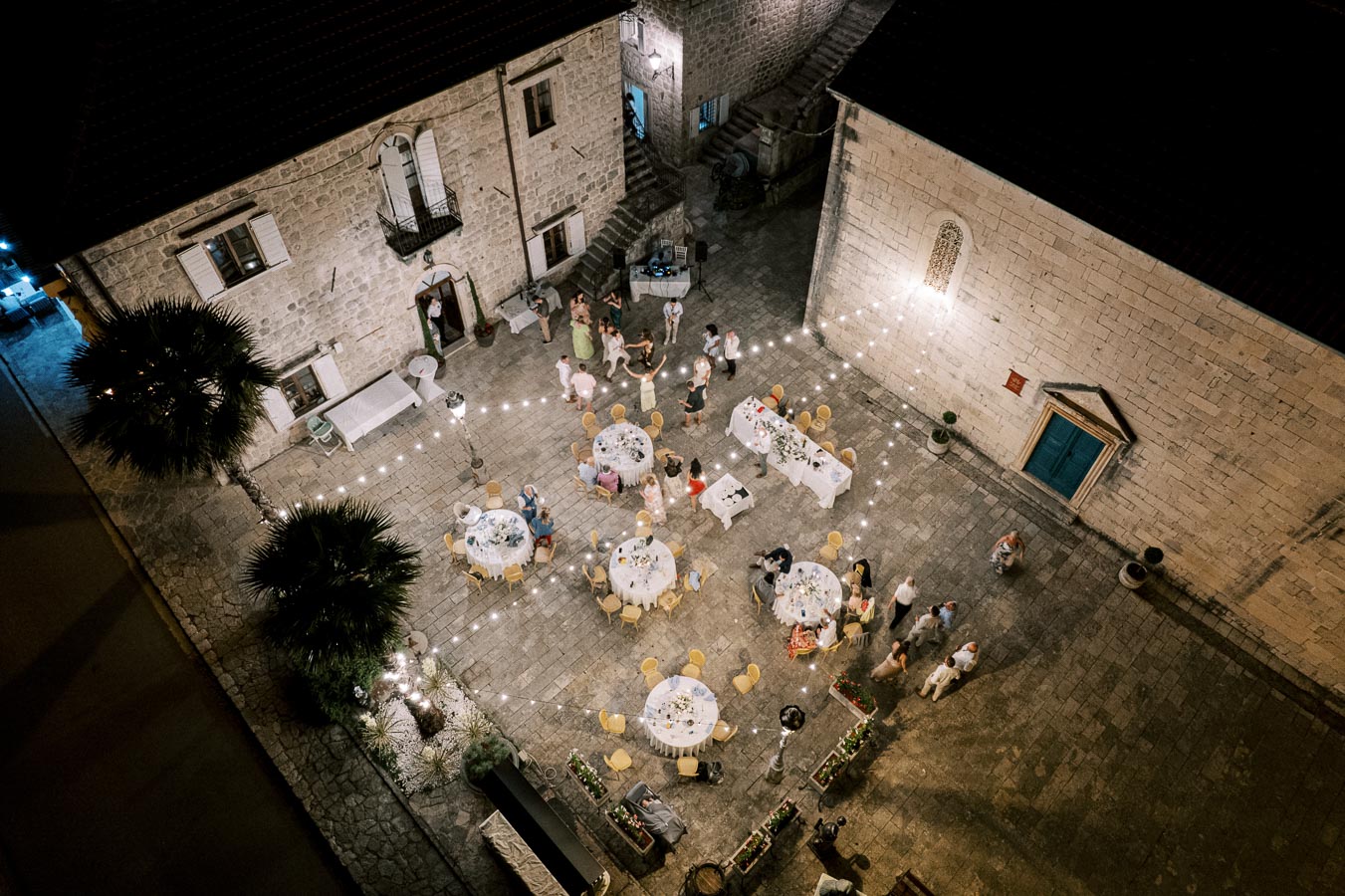 Guests enjoying an outdoor evening wedding reception in a charming stone courtyard, decorated with string lights and round tables.