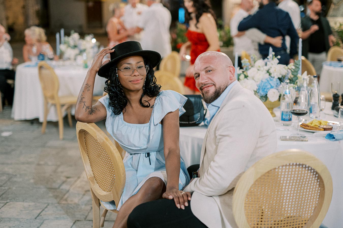 A couple seated at an outdoor event, woman in a light blue dress and black hat, man in a white blazer, surrounded by elegantly set tables with blue and white floral arrangements and guests in the background.