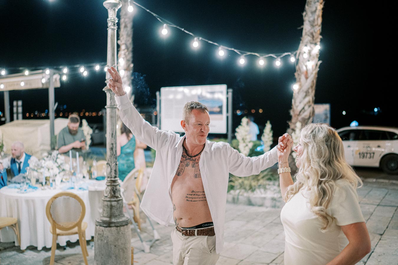 Outdoor evening wedding reception with guests dancing under string lights, featuring a man with an open shirt joyfully spinning a woman with long blonde hair on a stone patio.