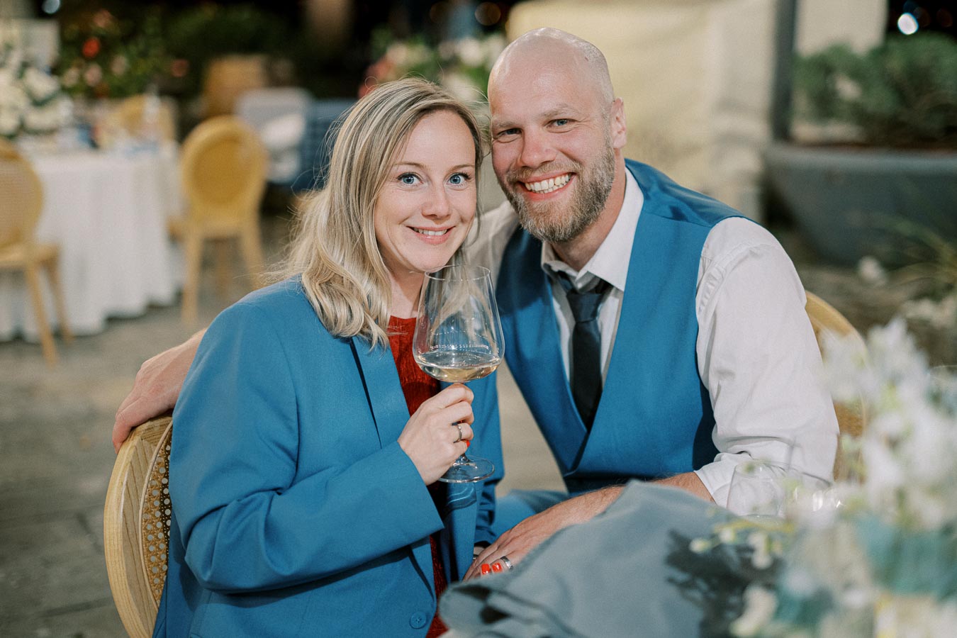 Smiling couple in matching blue outfits enjoying a glass of wine at an outdoor event with elegant decor in the background.