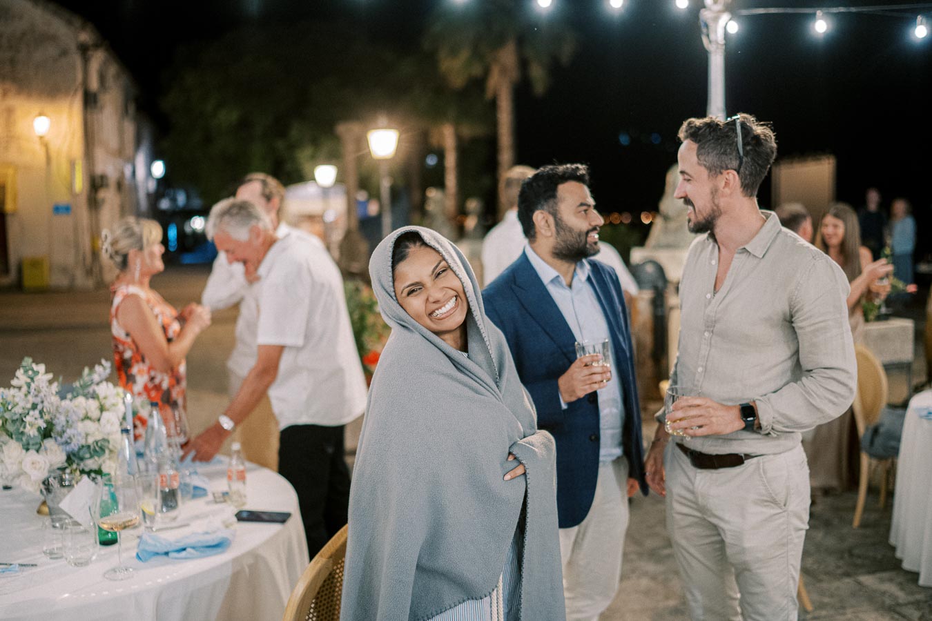 Group of people enjoying a lively evening outdoor gathering, with a smiling woman wrapped in a gray shawl in the foreground and others engaging in conversation in the background, surrounded by soft lighting and elegant decorations.