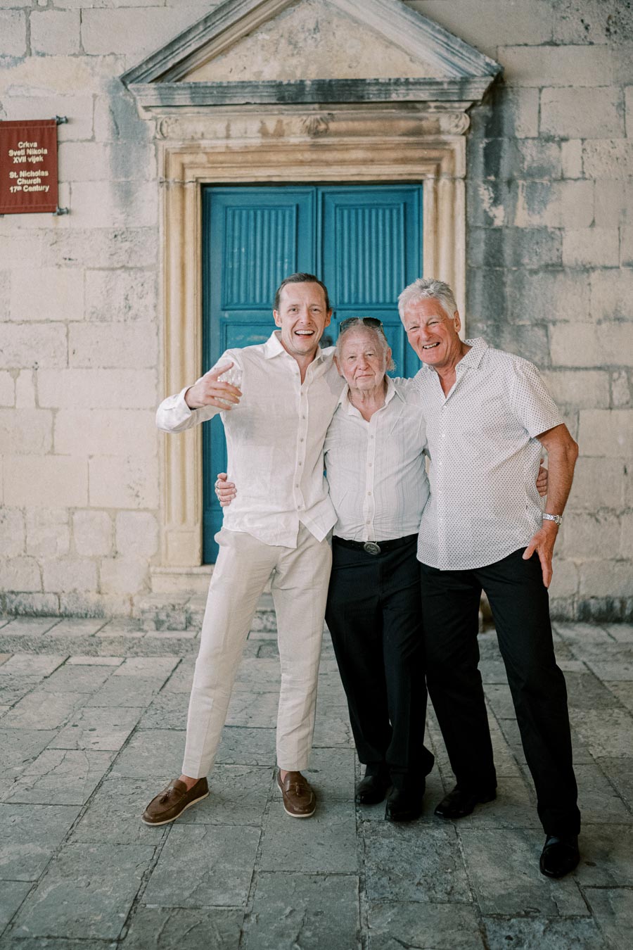 Three men in light-colored clothing pose happily in front of a historic stone building with a blue door.