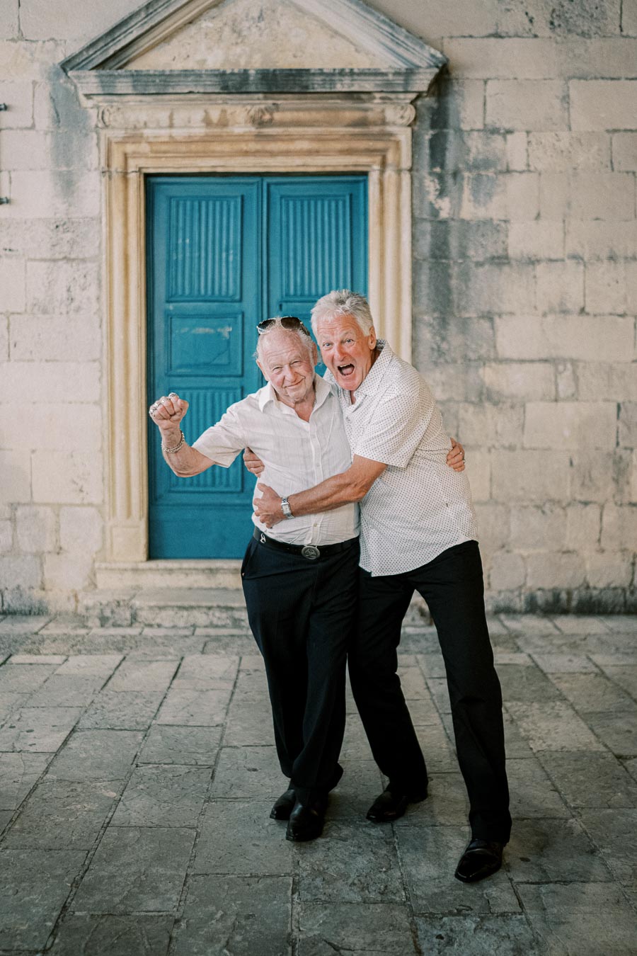 Two elderly men cheerfully embrace in front of an ornate blue door set in a rustic stone wall, showcasing friendship and joy.