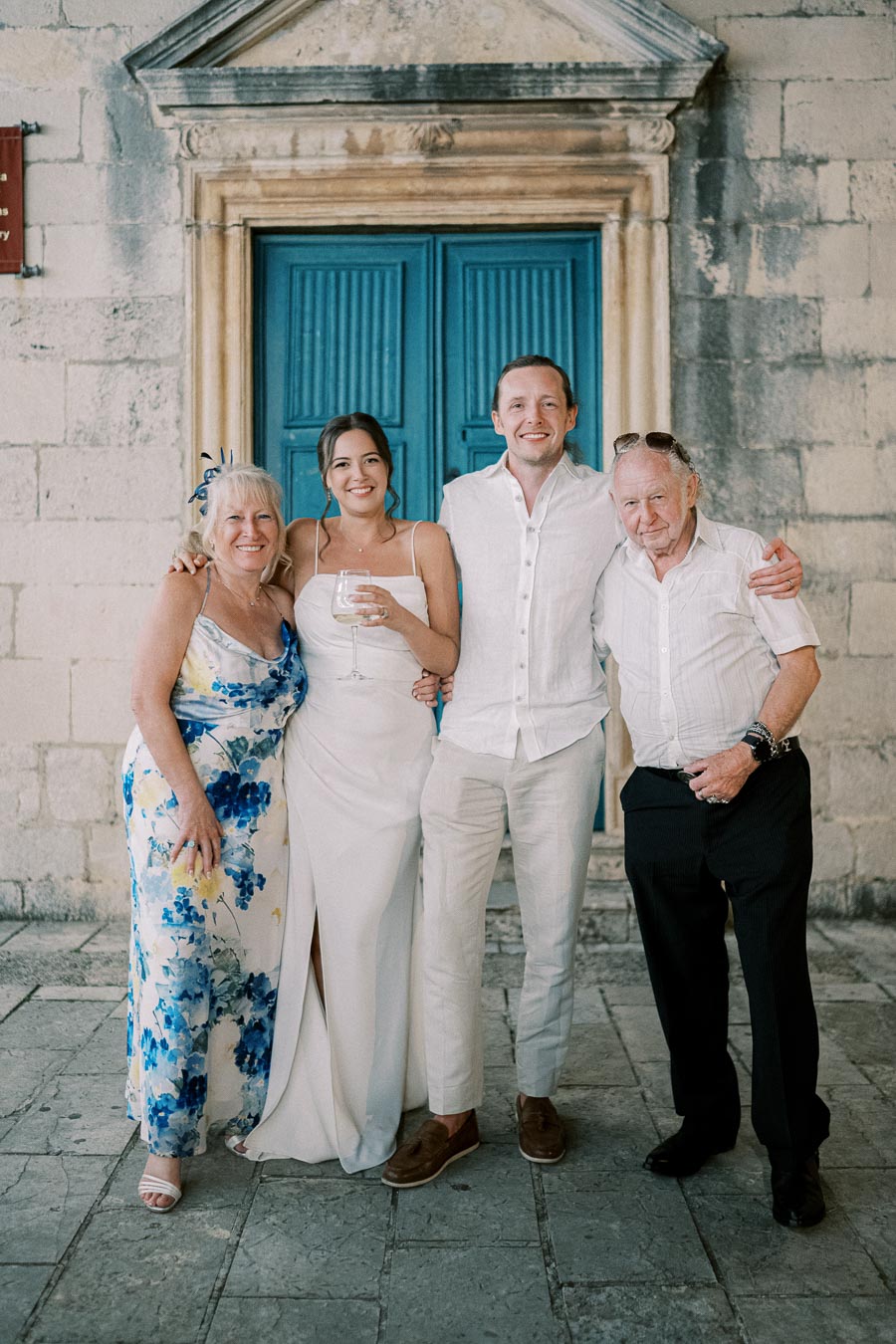 A group of four people smiling in front of a historic stone building with a blue door, dressed in formal attire for a special occasion.