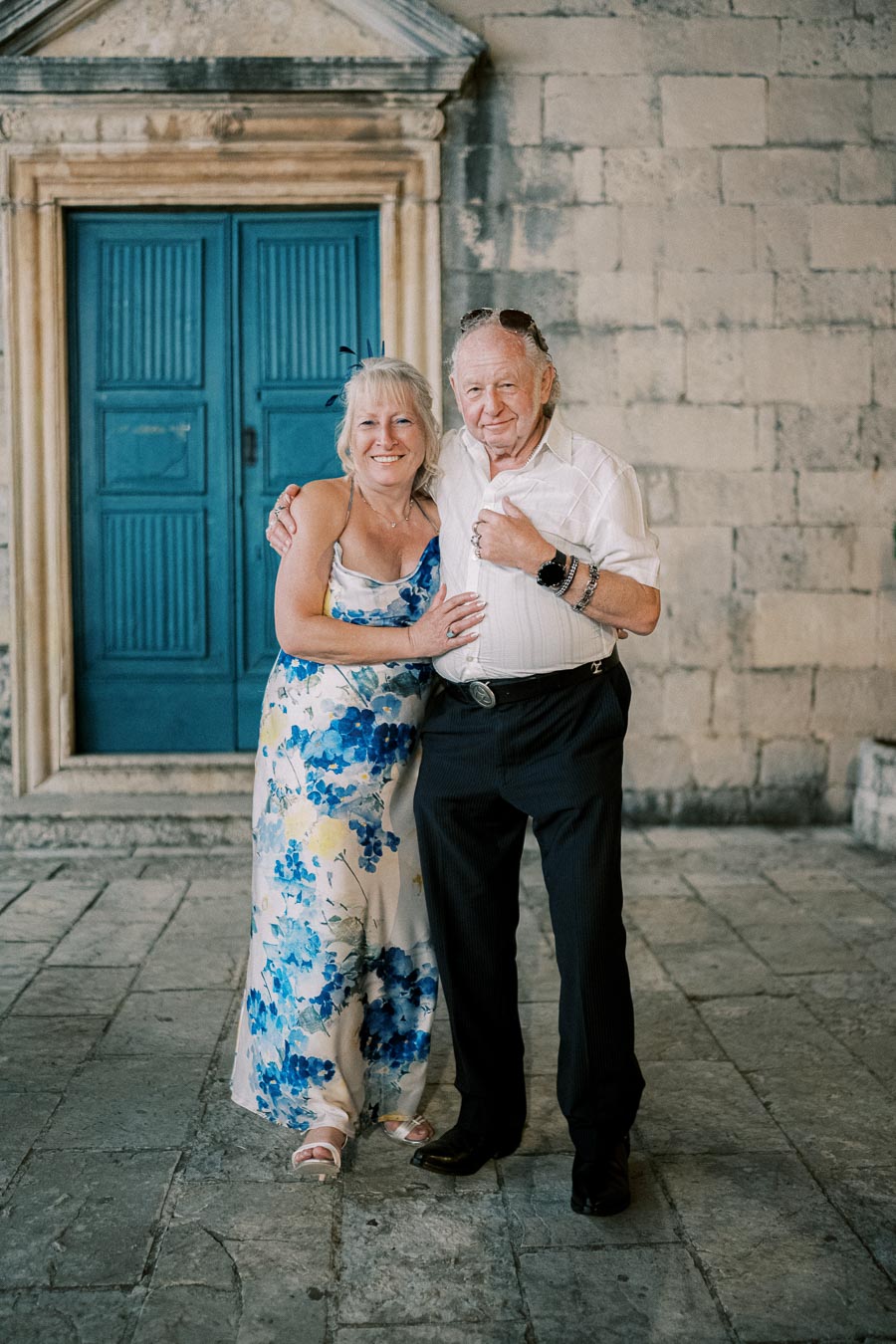 Elderly couple happily posing in front of an old stone building with a bright blue door, embracing and smiling, with the woman wearing a floral dress and the man in a white shirt and dark pants.