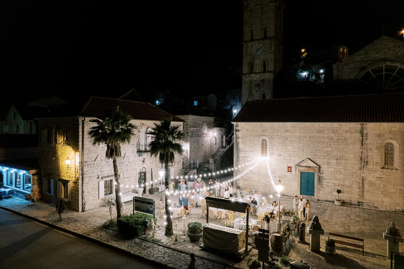 Nighttime outdoor event in a historic Mediterranean courtyard, featuring stone buildings with arched windows, palm trees, and twinkling string lights over dining tables set for an elegant gathering.
