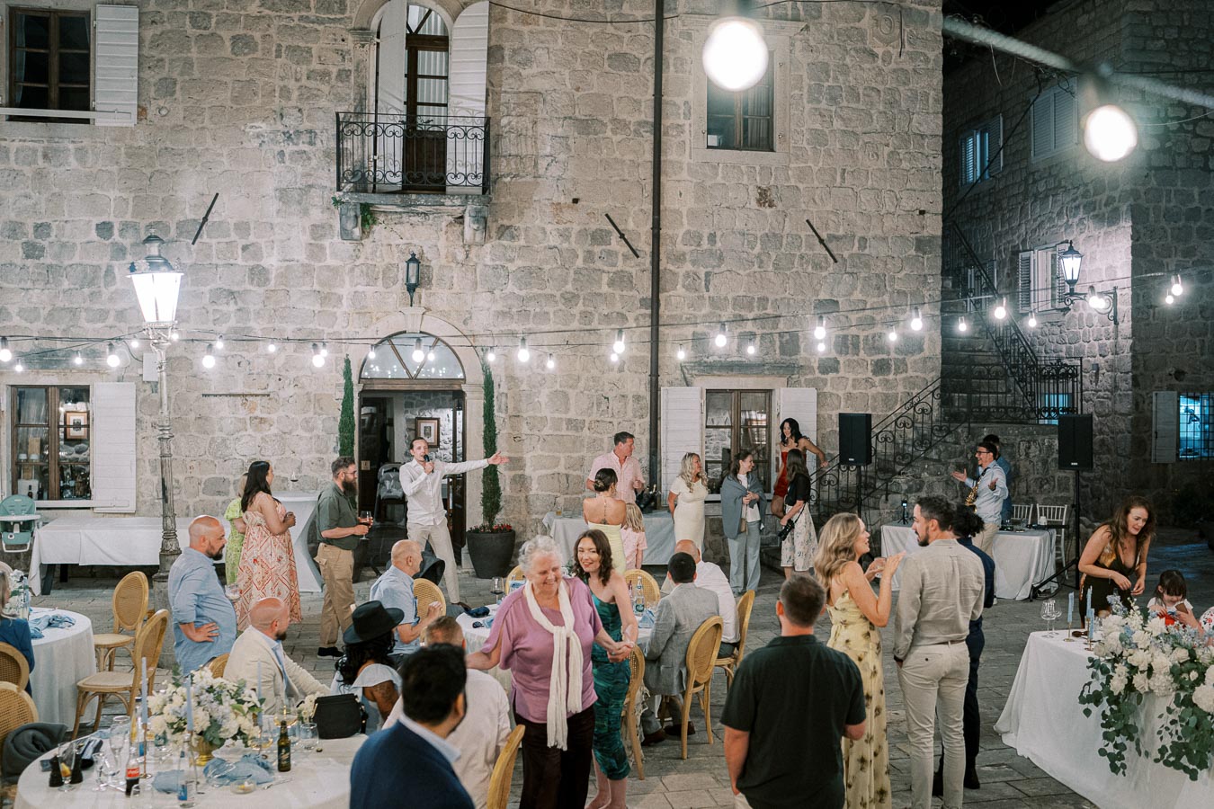 Outdoor evening wedding reception with guests socializing under string lights in a charming stone courtyard.