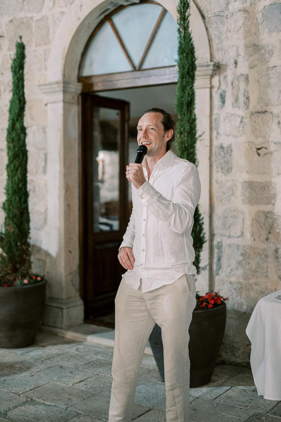 Man in white attire giving a speech with a microphone in front of a stone building entrance.