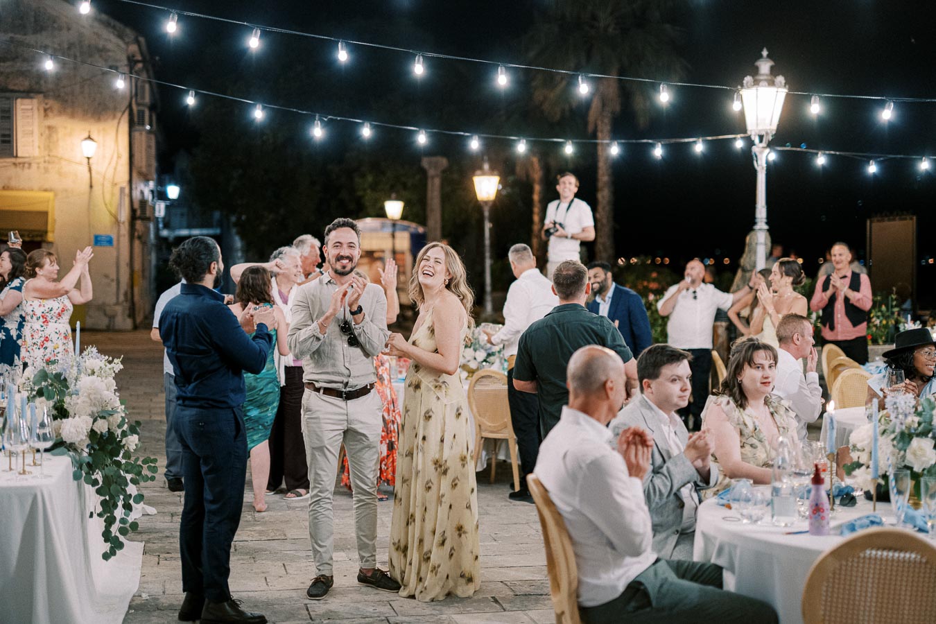 Outdoor wedding reception at night, featuring a lively group of guests applauding and celebrating under string lights, with tables elegantly decorated with flowers and candles.