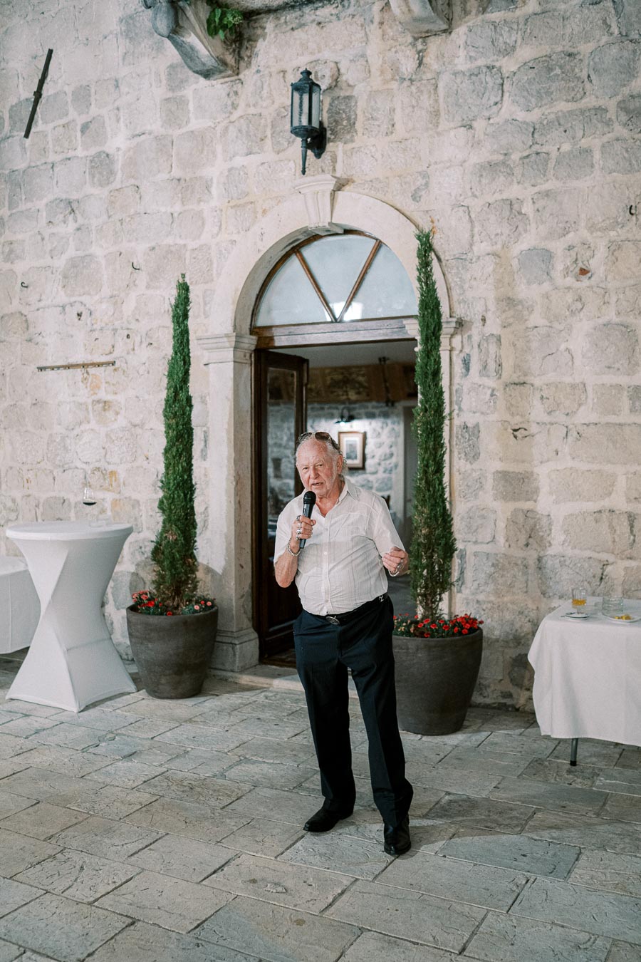 An elderly man holding a microphone gives a speech in front of a rustic stone building, decorated with potted plants and a small table with refreshments.