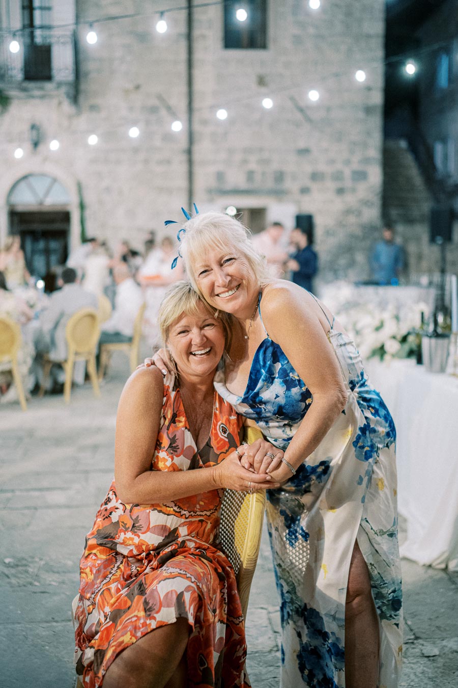 Two women smiling and embracing at an outdoor event, decorated with string lights and set against a rustic stone building backdrop. They are both wearing floral dresses and appear to be enjoying a festive gathering.