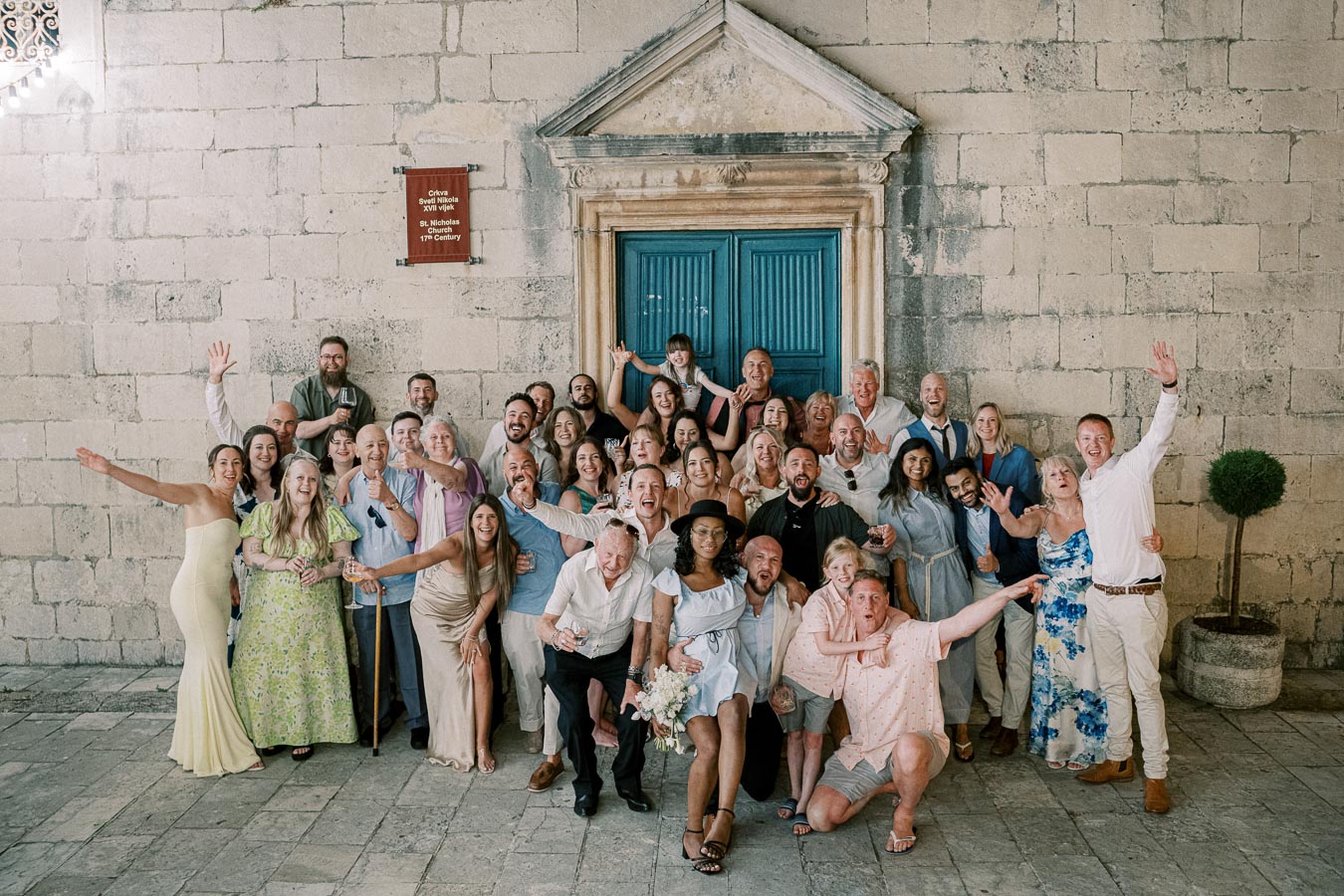 A large group of people celebrating together in front of a historic stone building with a blue door, capturing a joyful and festive moment at an outdoor event.