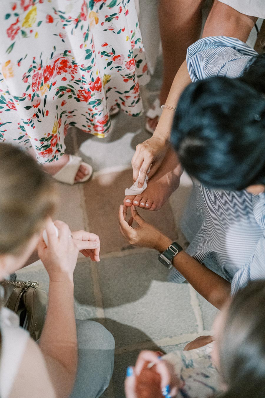 Group of people tending to a person's foot with a tissue, surrounded by floral dresses and tile flooring.